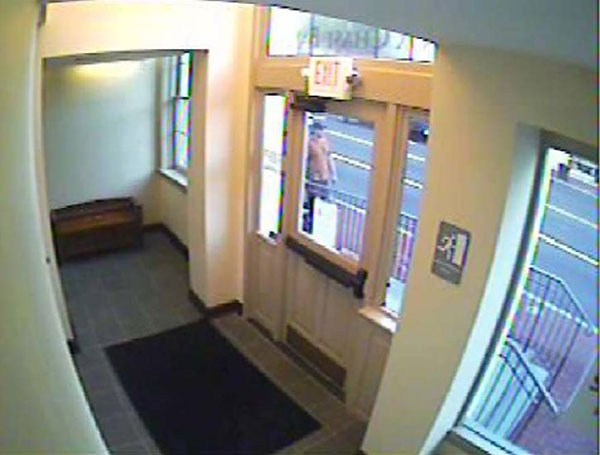 A surveilance camera photo of the front of the male suspect wearing an orange tshirt, dark pants, and a baseball cap. Photo was shot through a doorway window.