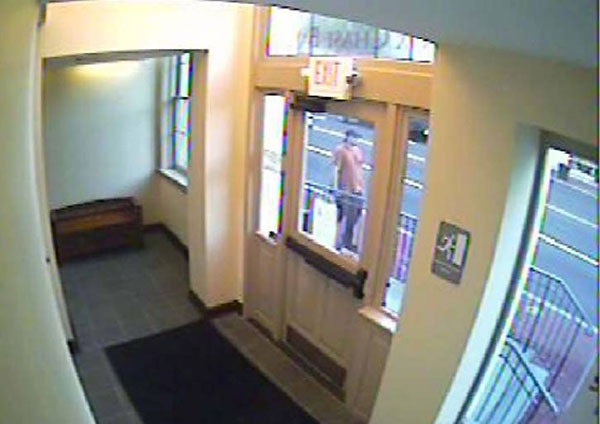 A surveilance camera photo of the front of the male suspect. Suspect is wearing an orange tshirt, dark pants, and a baseball cap.  Photo was shot through a doorway window.