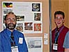 Teacher Mr. Moore stands to the left of a student poster project showing various satellite images of different locations as a student stands to the right of the project