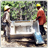 Two people wearing hard hats carry a broken-down refrigerator in a cleanup area.