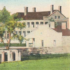 detail from a vintage postcard of officers quarters in distance and guard house on right with stone fort wall
