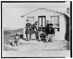 Group of five officers standing and seated in front of a house with a black boy, wearing cap, seated on the ground at their right