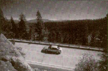 View from top of slope beyond to Lake Tahoe with one car on the road and trees and mountains in the background.