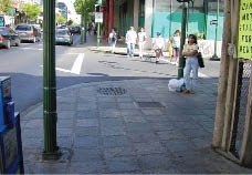 Case Study:  Two photo views of the same urban intersection show how an existing storm sewer inlet precludes the addition of a curb ramp within the crosswalk on one side of the crossing.