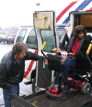 Photograph of a driver of a paratransit vehicle assisting a wheelchair-bound person to debark using a lift.