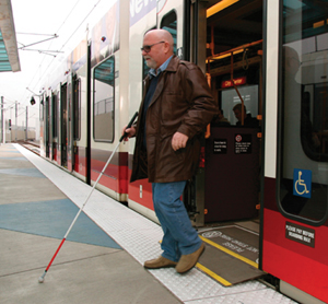 Photograph of a visually impaired person with a walking stick debarking from a commuter train equipped with a ramp covering the gap between the platform and the edge of the train's doorway.