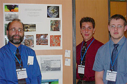 Teacher Mr. Moore stands to the left of a student poster project showing various satellite images of different locations as two students stand to the right of the project