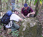 Two men in backpacks kneeling down to get a closer look at a large, moss-covered rock