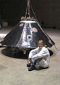 Jaime Dyk wears a white lab coat and safety goggles as she sits in front of one of the shells that covers the Mars rovers to protect them