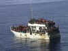 A side view small white vessel with large numbers of people on the deck and pilothouse.