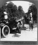 Man in horse and buggy greeting woman standing in automobile, while driver is underneath repairing it