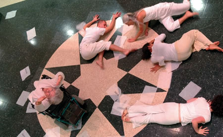 Shot of dancers taken from above.  Some are laying down on the floor while a female on a wheelchair stretches her arms toward the camera
