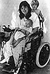 Photo: A mother sits in a wheelchair smiling, with a baby in her lap and a small boy standing up behind her on the back of her chair.