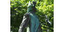 A green statue stands among trees, it has a soldiers uniform on, with a cocked hat, and a sword.