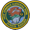Junior Ranger patch is fully embroidered with an arrowhead shape filled with a raccoon in the foreground, pine trees on either side with a mountain, mockingbird and butterfly.
