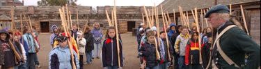 Children stand at attention in rows as straigh as trees, with wooden muskets in a parade ground, waiting for a continental soldier's orders.