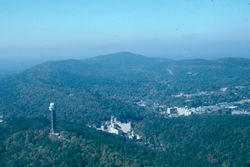 aerial view of Hot Springs Mountain with the Mountain Tower on the lower left, West Mountain and Music Mountain above the tower and the valley with downtown buildings showing on the right. Photo has a blue hue.
