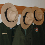 Three National Park Service straw flat hats and green jackets hung up in a row.