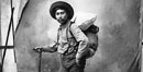 Historic photo of Native Tlingit packer carring a pack of goods on his back, wearing Western gear