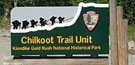 Chilkoot trailhead sign showing the National Park Service arrowhead logo and an outline of people with loads climbing up a steep, snowy pass