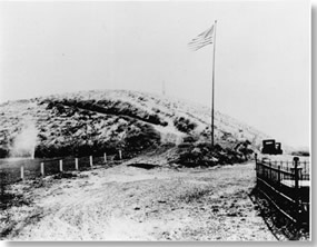 Historic photo of hill. Large flag pole at base of hill flying American flag. Two cars parked at base of hill.