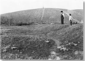 Two men standing on small rise looking down at lower land. No water in depression.