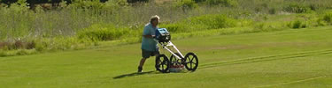 Archeologist is pushing a cart across park grounds. On the cart is an antenna that is part of the ground-penetrating radar survey system.