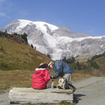 One day hiker sits on bench while another hiker stands next to her as they plan their next hike.