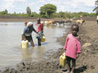 Woman and children fill water containers on muddy banks of body of water. Photo courtesy of Photoshare.