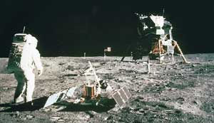 The first people on the moon were U.S. astronauts Neil A. Armstrong, who took this picture, and Buzz Aldrin, who is pictured next to a seismograph. A television camera and a United States flag are in the background.