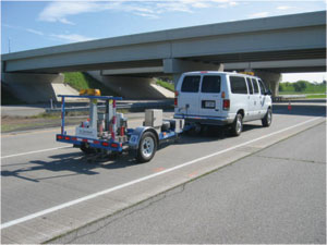 Photograph. A van is towing falling-weight deflectometer testing equipment behind it over a section of newly laid concrete pavement in a Minnesota project.