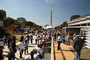 Photo of large groups of people visiting a variety of modern-looking houses lining the National Mall. The Washington Monument rises in the background against a clear blue sky.