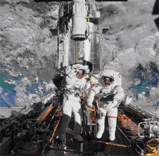 Two astronauts in white suits standing outside on the shuttle in space wave to the cameraman inside the space shuttle