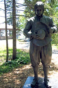 A bronze statue of a French settler holding his hat sits in the trees along an interpretive trail with a shelter in the background.