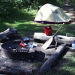 A light colored tent behind a fire ring with campstove on the grill.  Two logs are pulled up for seating