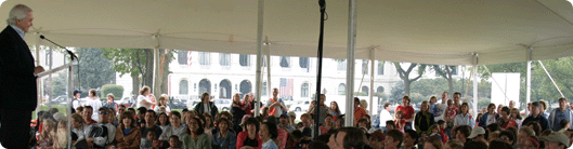 Large group of people under a tent, watching a man speak at a podium.