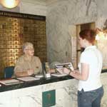color photo of volunteer Bettie Gardner orienting visitor to the visitor center, standing at the original Fordyce Bathhouse business counter with marble top and brass lock boxes behind