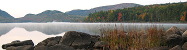 Glassy lake with rocks and grasses in foreground, fall colors in background
