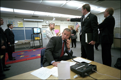 As Director of Communications Dan Bartlett points to news footage of the World Trade Center Towers burning, President George W. Bush gathers information about the attack at Emma E. Booker Elementary School in Sarasota, Fla., Sept. 11, 2001. Also photographed are Director of White House Situation Room, National Security Council, Deborah Loewer (directly behind the President) and Senior Advisor Karl Rove (right).