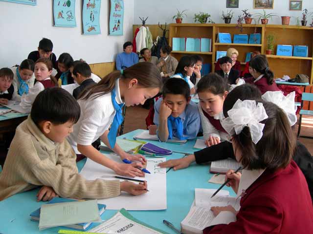 A photograph of children sitting at a table with paper, pens, and books.  