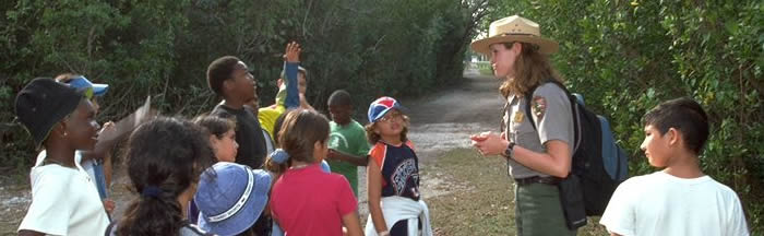 A ranger answers questions asked by a group of school children as they stand at a wide spot along a trail through trees and brush.