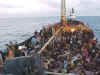 The view from the pilot house of a migrant vessel with large numbers of people on deck.  Off in the distance is a Coast Guard High Endurance Cutter.