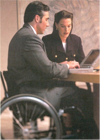 Picture of a woman having a discussion with a man sitting in a wheelchair as he types on a laptiop 