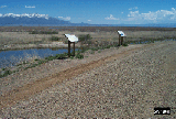 photograph of the gravel auto tour road at the Alamosa National Wildlife Refuge, Alamosa, Colorado, showing two interpretive signs, a marsh in the foreground, high desert shrub vegetation behind the marsh and the snow covered mountains in the background.