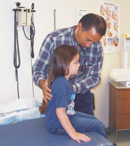 A young female patient receives care from a doctor at Carlsbad Family Health Center. She is sitting on the examination table while the doctor holds a stethoscope to her chest. 