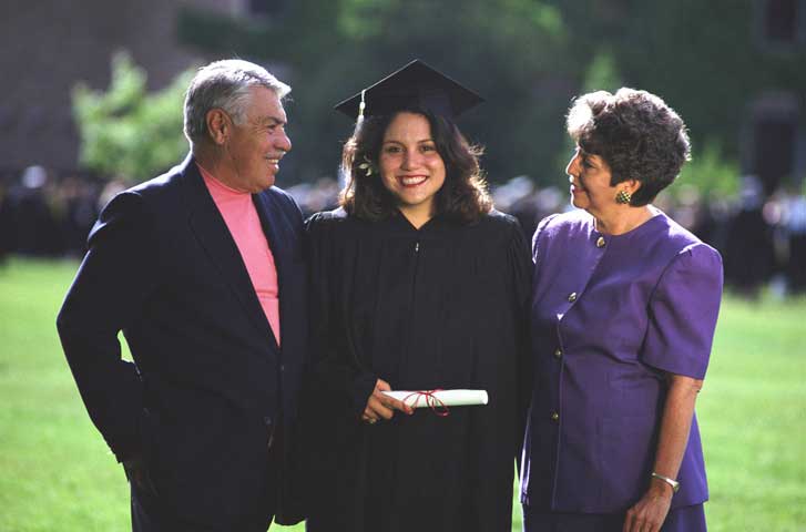 This is a photo of a smiling young woman in her graduate cap and gown attire with her parents proudly standing next to her.  