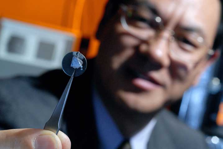 The image shows a researcher holding a small metallic disk toward the camera using sharp tweezers. The mechanism on the disk is too small to easily discern its design, eve in close-up.