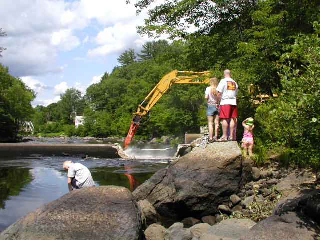 Photo of the Henniker dam in New Hampshire being removed by a backhoe. People are standing on rocks at the water’s edge watching the removal. 