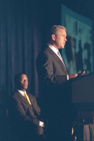 A profile picture of President Bush standing at a podium speaking while Rep. J.C. Watts, seated behind him, looks on.