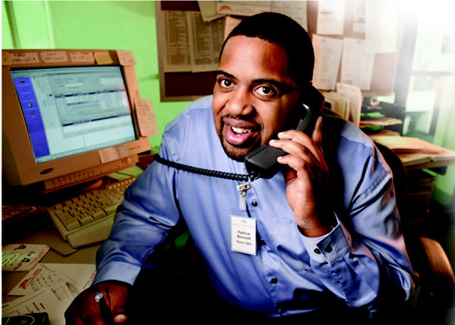 This Photo shows a man sitting in front of a computer and smiling as he talks on the telephone.